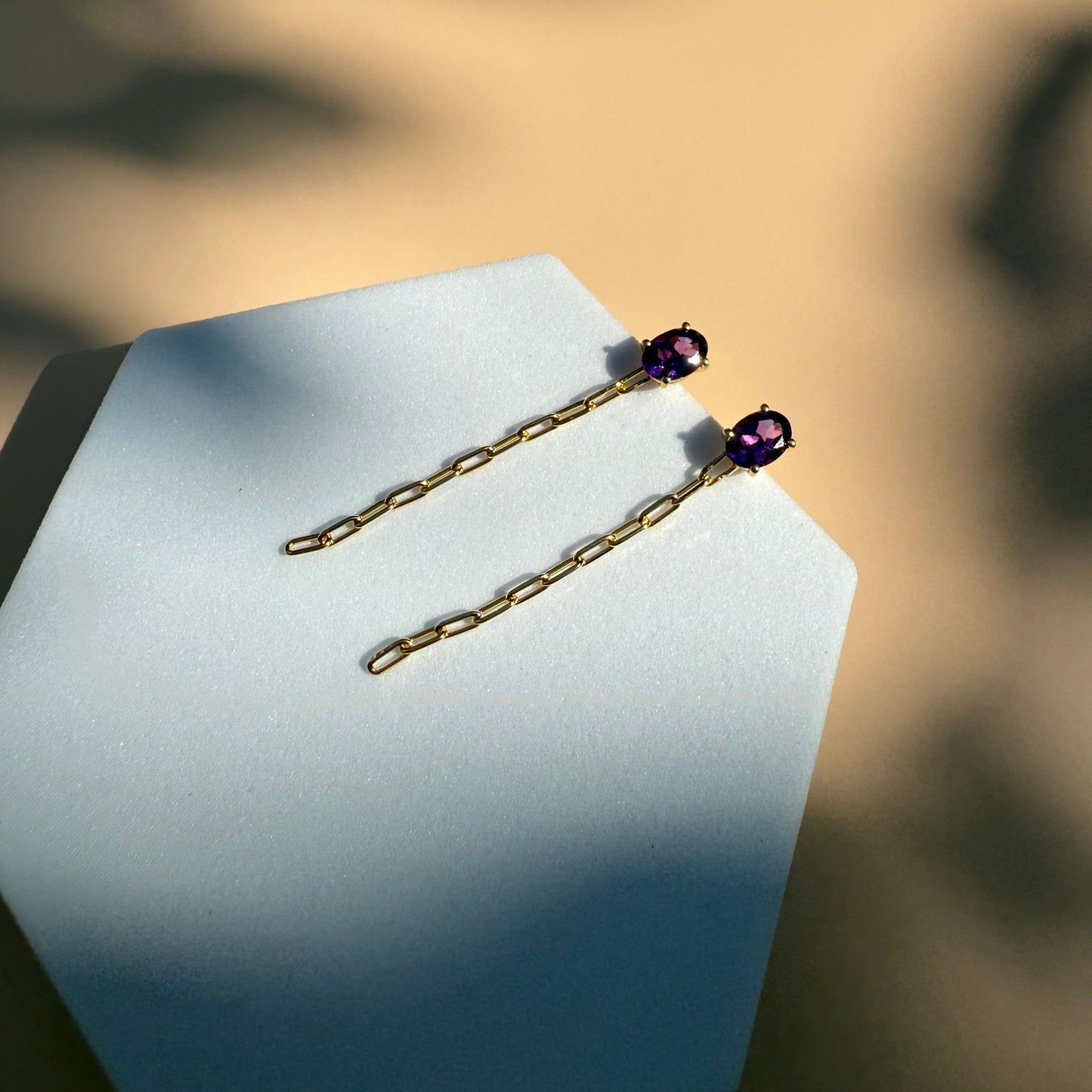 Gold amethyst earrings with dangling chain on a white hexagon 